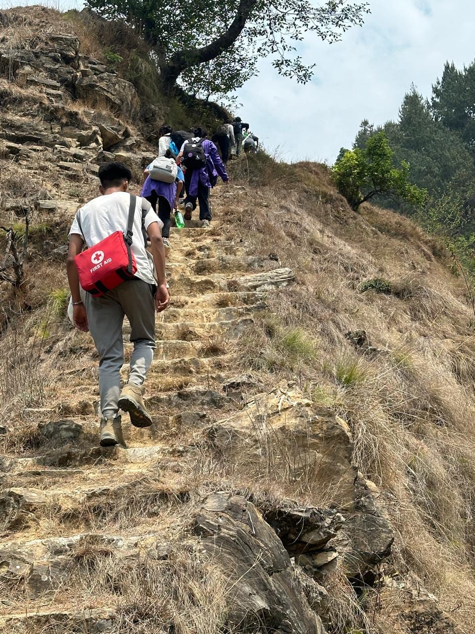 Students climbing a rocky hillside path on the way to Balthali, one carrying a red first-aid bag