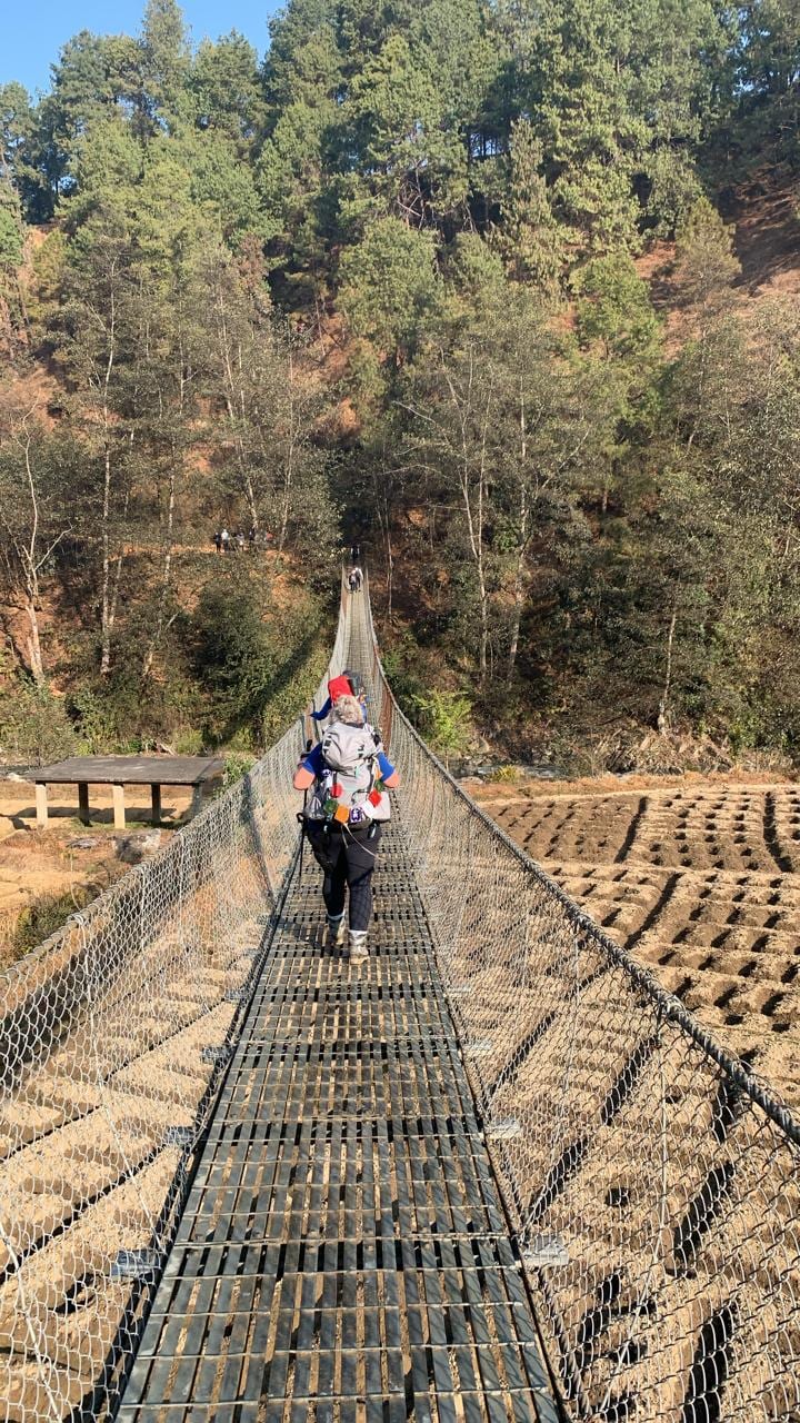 A walker with a backpack crossing a long steel suspension bridge over the river on the way to Balthali