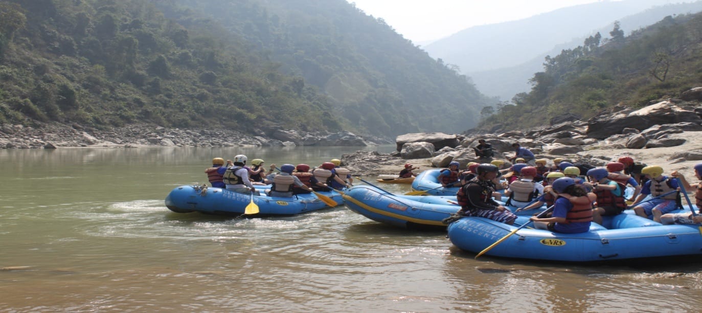 Blue rafts full of paddlers in helmets and life jackets gathered on a wide river below forested hills
